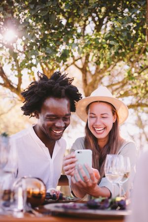 A man and woman laughing at something on the phone screen over lunch outdoors