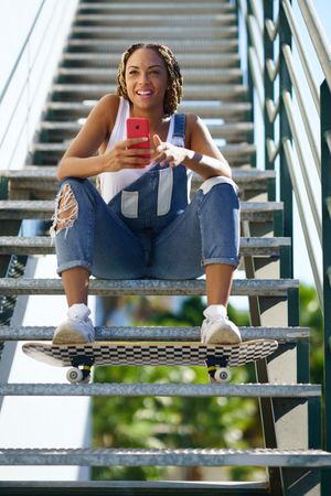 Happy female checking phone on stairs with skateboard