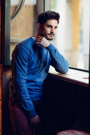 Thoughtful man in blue sweater lost in thought near a window in a modern pub