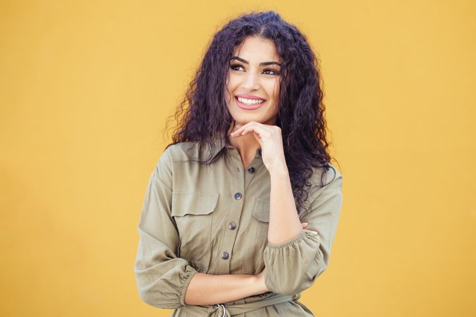 Female in army green jumpsuit smiling in front of mustard wall resting chin on her hand
