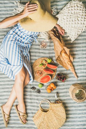 Woman in hat on picnic blanket with  thatched bag with glass and bottle of rose, baguette