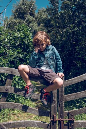 Pensive preteen boy sitting on wooden fence in sunny day