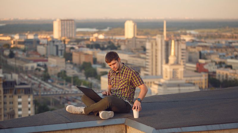 Man sitting crossed legged on roof over looking city with laptop and takeaway drink