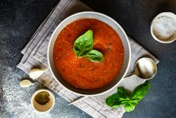 Top view of grey bowl of gazpacho soup with basil leaves on napkin served with seasoning