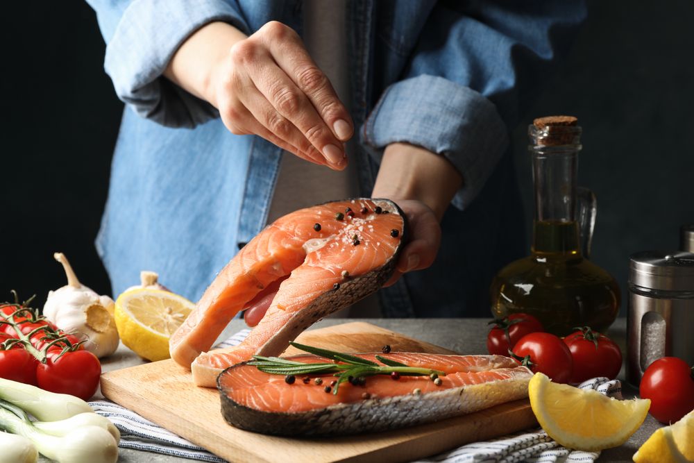 Woman sprinkles salt on salmon meat. Cooking salmon on gray table