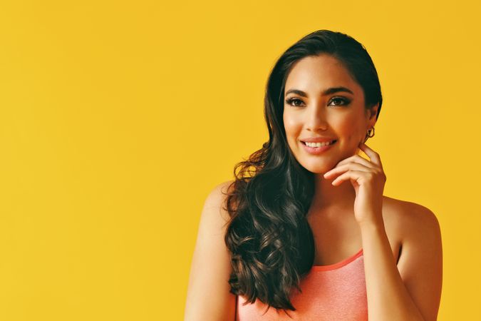 Headshot of smiling Hispanic woman looking away in yellow room, with hand up to her cheek