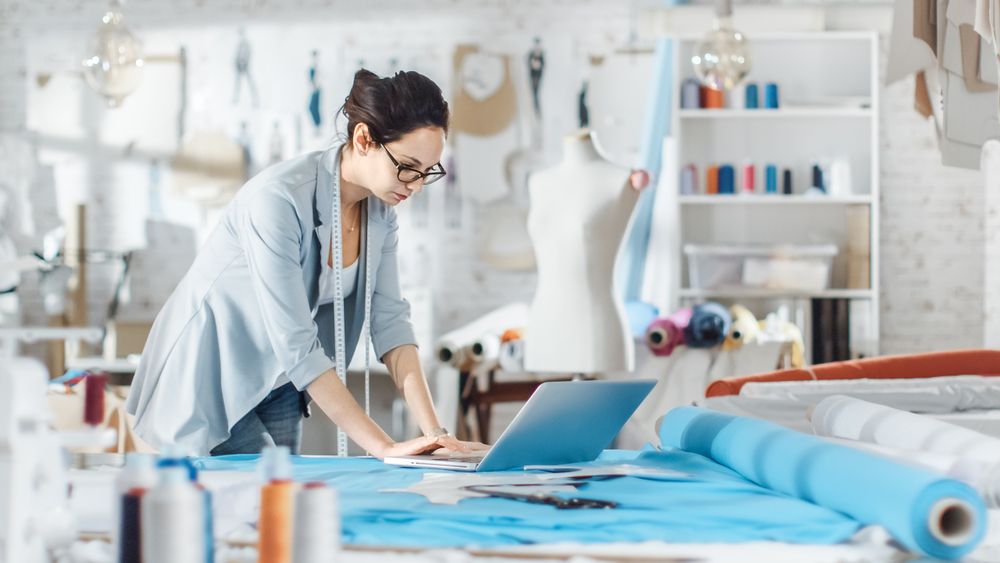 Female Designer Uses Laptop on Her Desk that is Covered with Various Fabrics and Sewing Items.