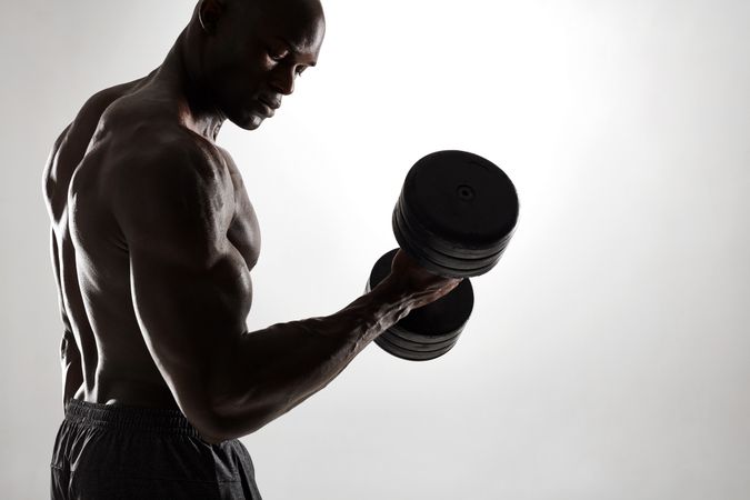 Side view of healthy muscular young man working out with dumbbells against grey background