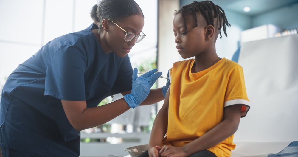 Female Black Nurse Is Performing Injection. Professional Woman Talks To Worried Kid And Calms Him Down