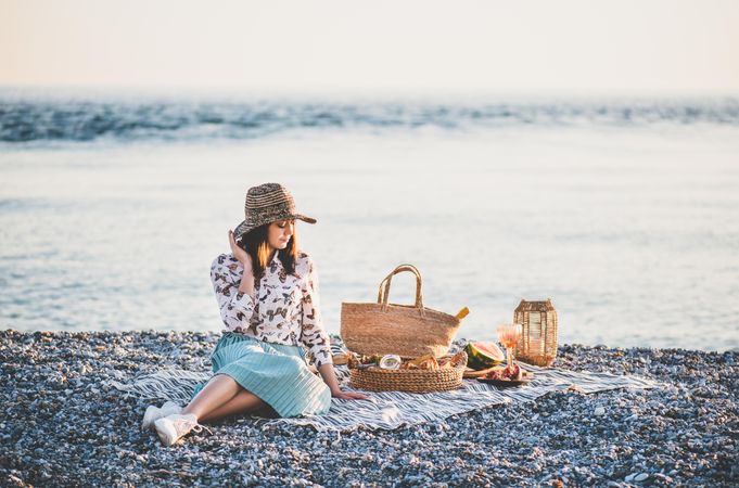 Young woman in hat sitting by the ocean with a picnic, watermelon, and wine