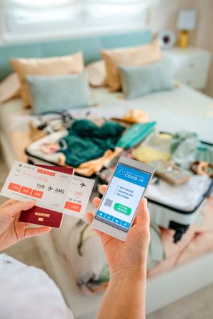 Hands of woman holding smartphone with covid health passport while preparing the suitcase
