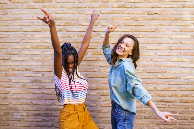 Two female friends dancing with their arms up celebrating in front of a brick wall