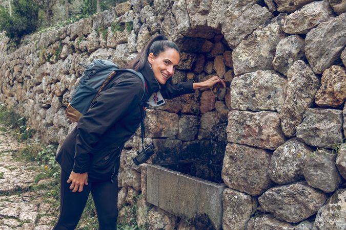 Positive female hiker standing near stone wall with flowing spri