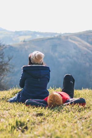 Back view of two people sitting on grass field facing mountainous landform