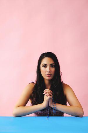 Hispanic woman praying with rosary beads in pink room, vertical