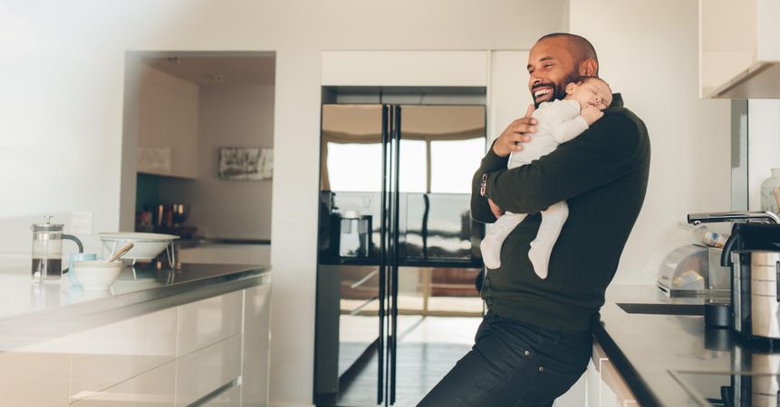 Happy man carrying his newborn baby boy in arms in kitchen