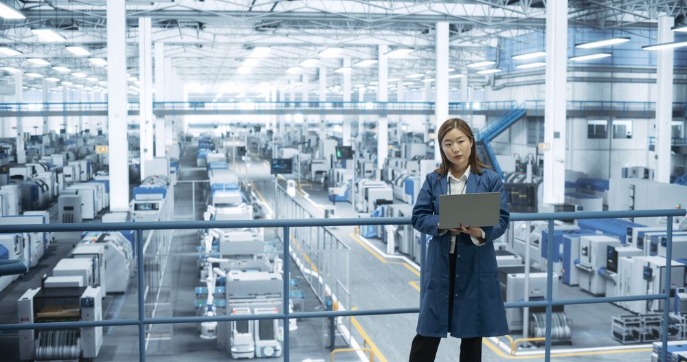 Shot in an Electronics Factory. Japanese Project Manager Working on Laptop at a Futuristic Industrial Manufacture with AI Robotic Arms. Specialist Monitoring Production on a Computer