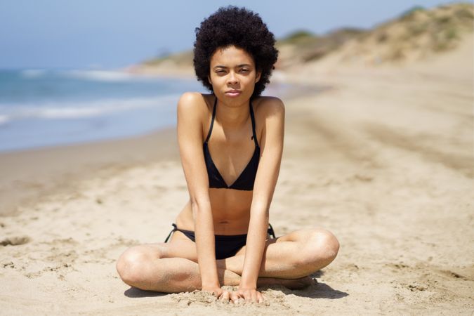 Serious female in bikini sitting crossed legged in the sand looking at camera