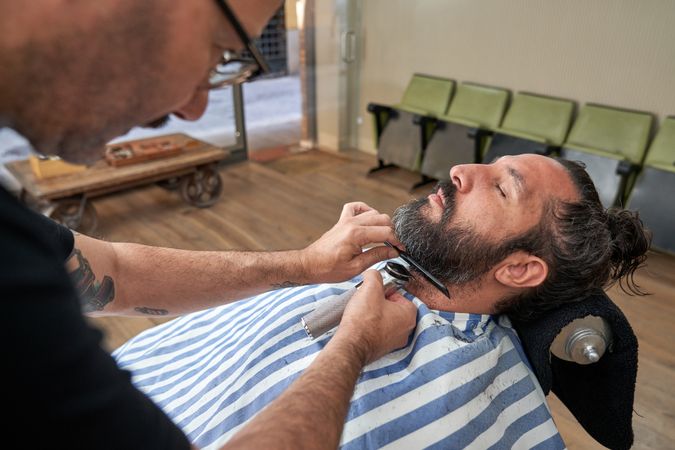 Crop barber trimming beard of customer in salon