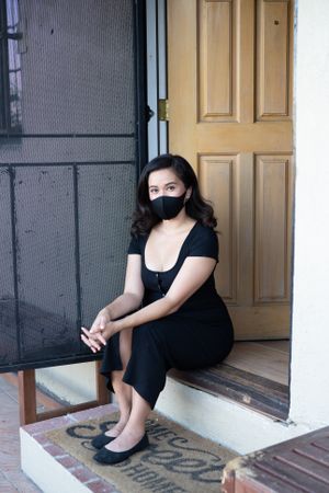 Woman sitting on front door steps of her house wearing mask and looking at camera