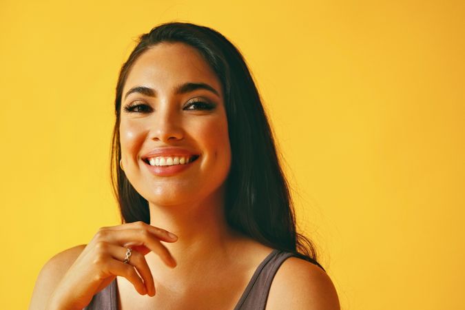 Happy confident Hispanic woman looking at camera in yellow room with her hand to her chin