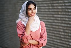 Female in headscarf and pink trench coat standing confidently with with one hand to her chin