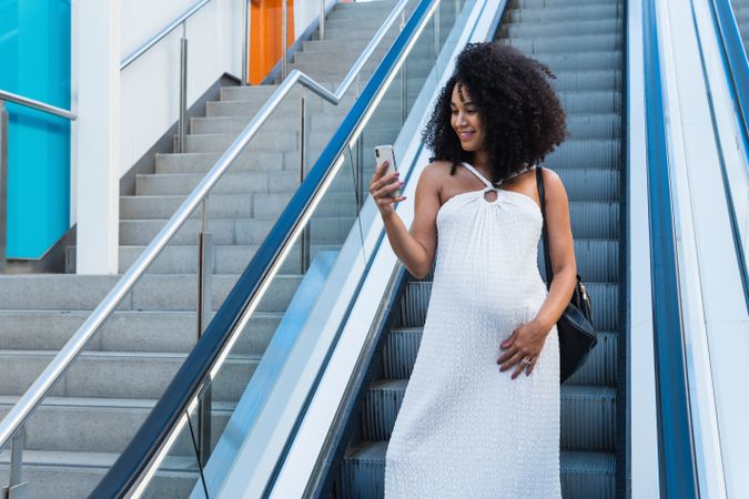 Pregnant Black woman browsing smartphone on escalator