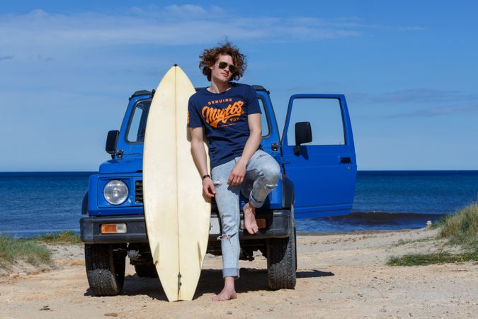 Guy with surfboard barefoot leaning on car on beach