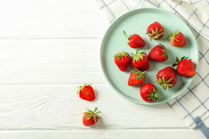 Plate with fresh strawberries and kitchen towel on bright wooden background, top view and space for text