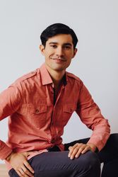 Hispanic male sitting in grey studio smiling in pink shirt
