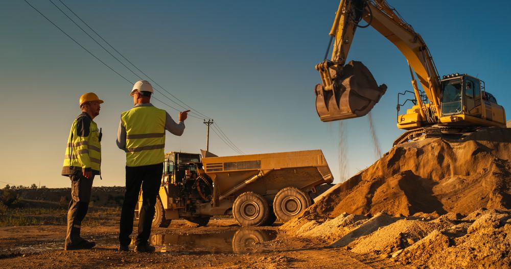 Male Real Estate Investor And Project Manager Talking On Construction Site Of Apartment Block. Colleagues Discussing Building Progress. Excavator Loading Sand In Industrial Truck On Warm Day