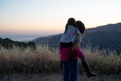 Back view of mom giving piggy back to her daughter and looking toward view of mountains and ocean
