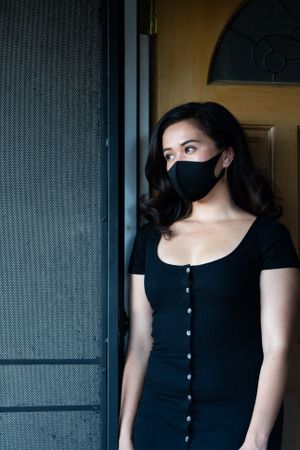 Portrait of woman looking out of her doorway wearing PPE mask and dress