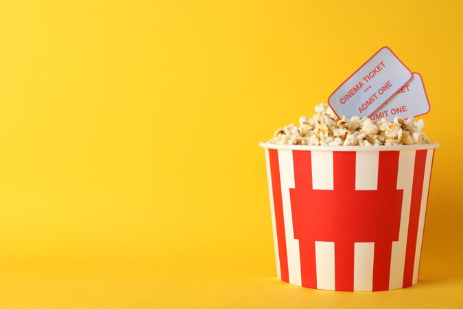 Striped bucket with popcorn and tickets on yellow background, copy space