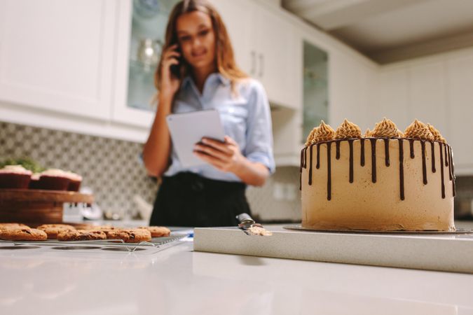 Young woman using cell phone holding tablet with cake on counter in kitchen