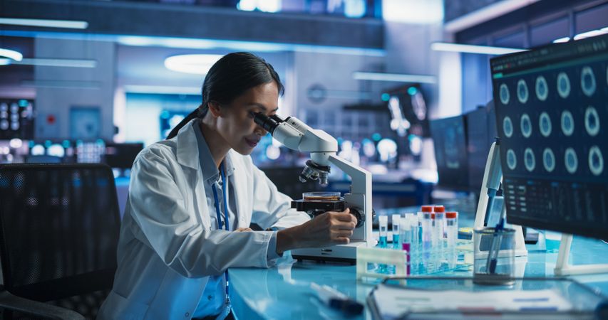 Medicine Development Laboratory: Asian Female Scientist Using Microscope, Analyzes Petri Dish Sample.