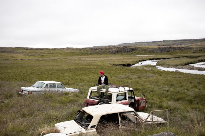 Man sitting on top of abandon cars in a field