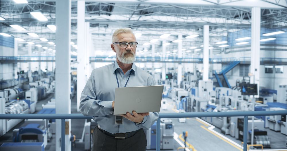 Experienced Male Engineer Standing on a Platform, Using Laptop Computer and Overlooking Production at a Modern Automated Electronics Manufacture with AI Support. Factory Executive at Work