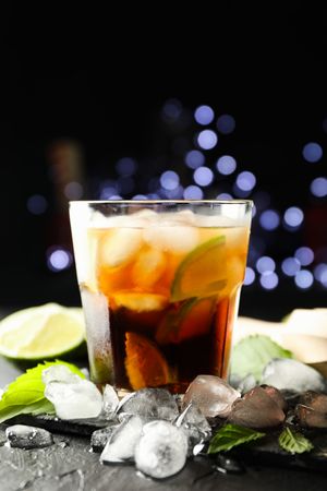 Glass with cola and lime slices, juicer, ice, mint against blurred lights background