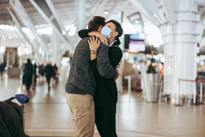 Woman in face mask giving welcome hug to man at airport arrival gate