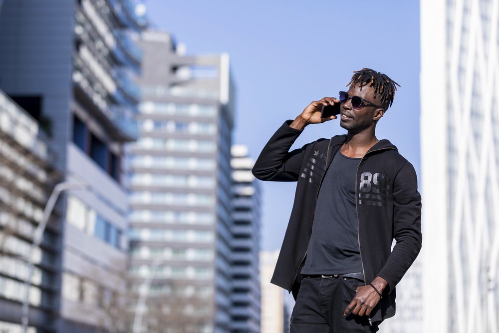 Black man with sunglasses standing against cityscape on the street while using a mobile phone in sunny day