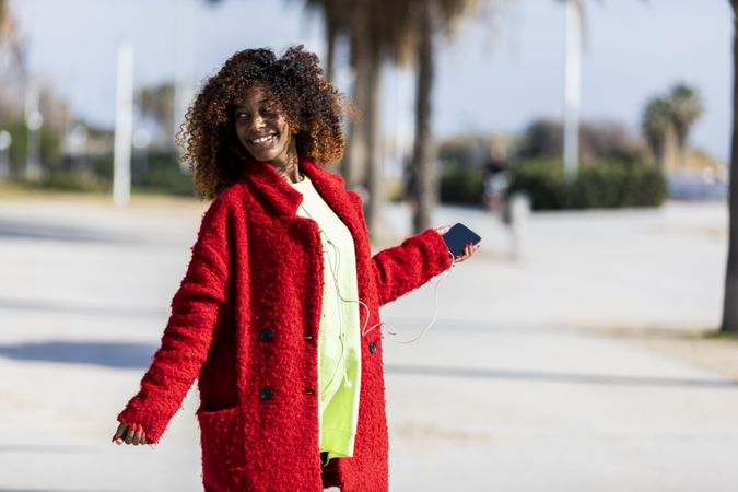 Happy female in colorful clothes listening to music on phone standing on street with palm trees