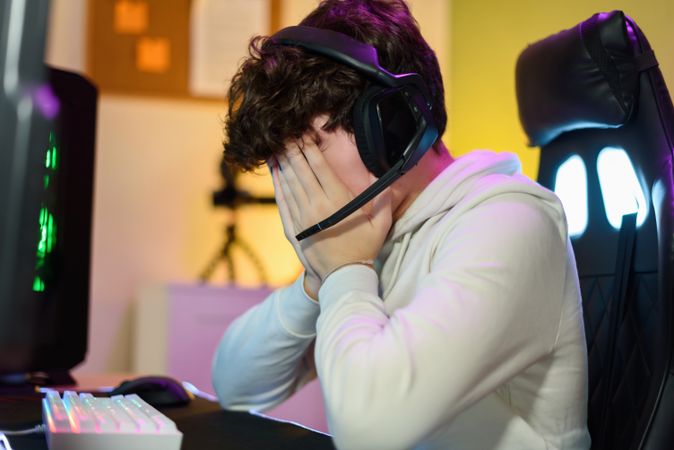 Anonymous young man sitting and covering face with hands near gaming PC