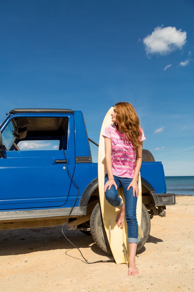 Woman leaning on surfboard near car at seaside