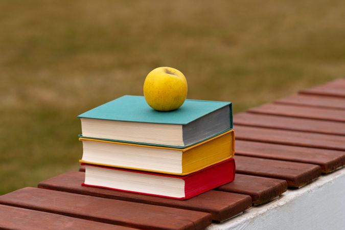 Stack of colorful books with apple resting on bench.