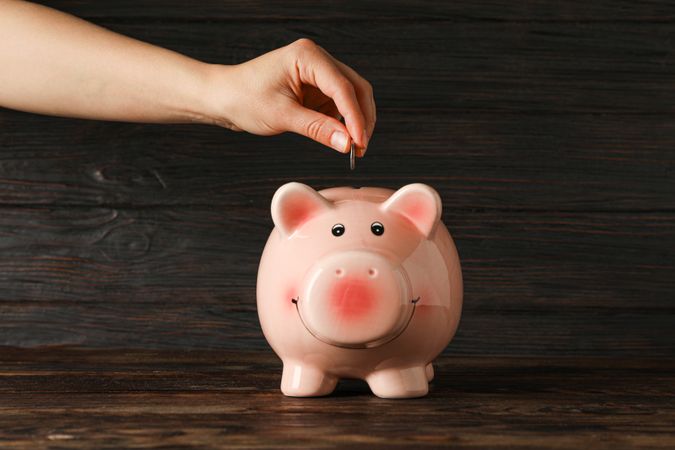 Woman's hand puts a coin in the happy piggy bank on wood table against wooden background, space for text. Finance, saving money