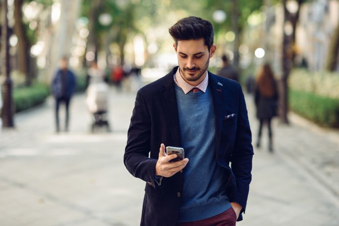 Attractive man in the street wearing elegant suit looking down at smartphone in his hand
