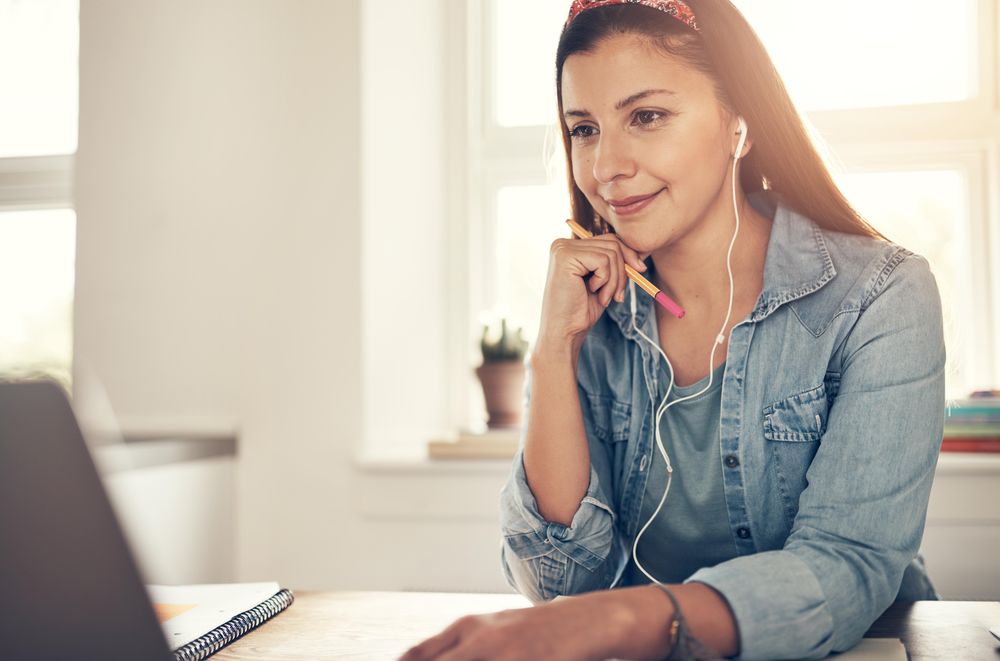 Contemplative woman reading on laptop in home office with earbuds and pencil