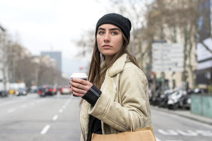 Portrait of female in hat crossing the street, with takeaway coffee and shopping bag on cold day