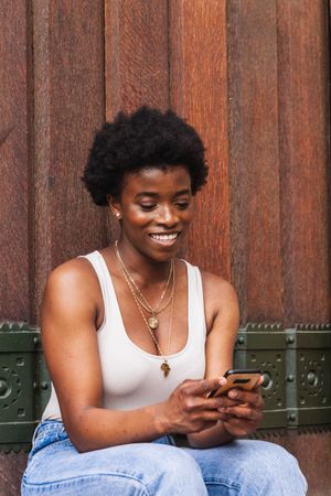 Smiling Black woman using smartphone sitting near building